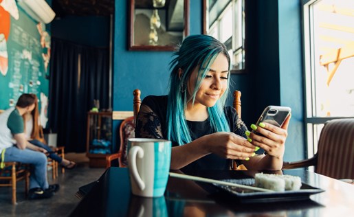 A woman with blue hair on her phone while eating sushi at a restaurant.